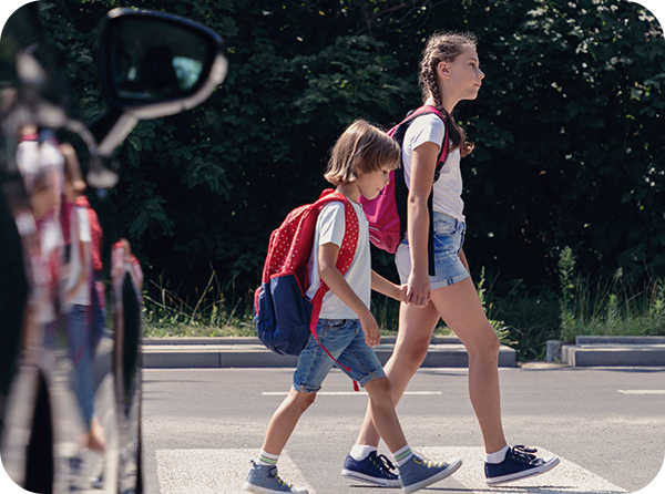 Photo of middle school-aged girl holding hands and walking across a crosswalk with an elementary-aged girl while a car waits to go
