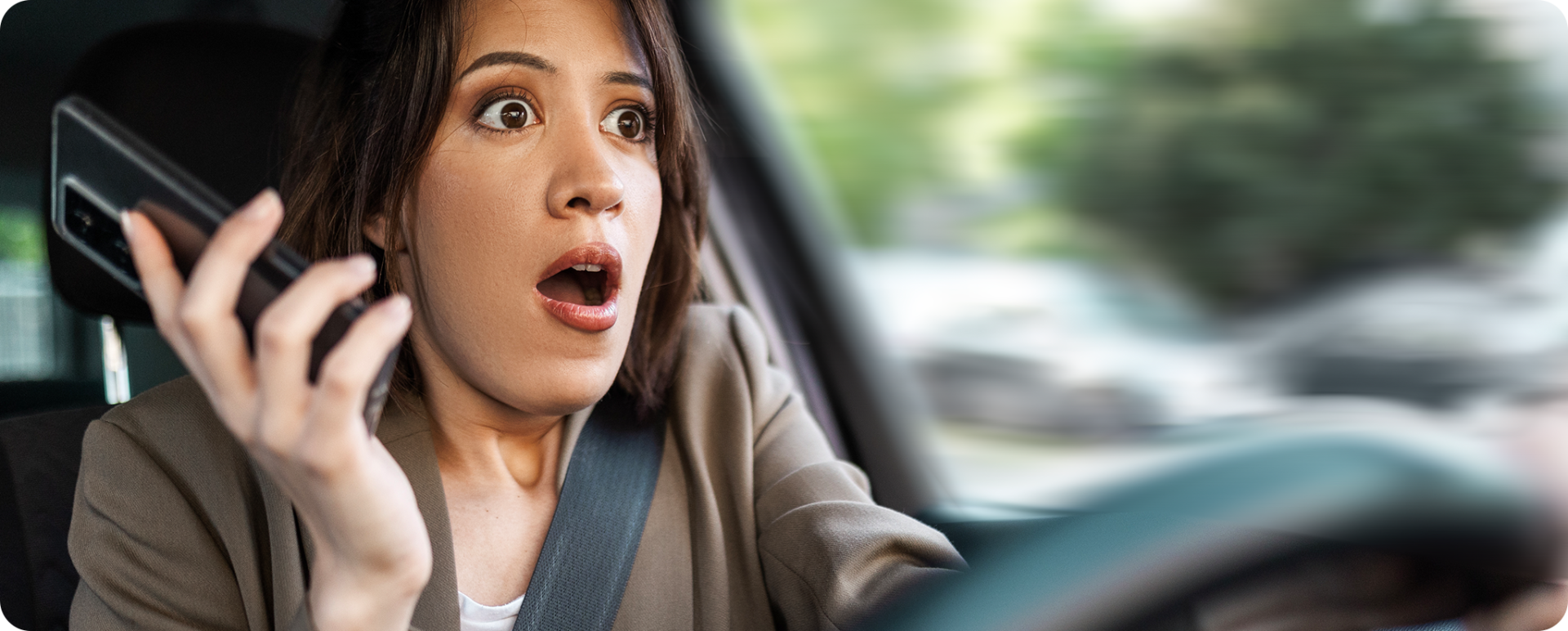 Photo of woman holding a phone and appearing shocked as she looks out her windshield, indicating she may not have been paying attention and has an unexpected road hazard in front of her