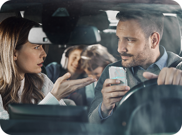 Photo of male driver holding a phone and listening to his wife in the passenger's seat who has her hand up in frustration. Two young kids with headphones on are in the back seat.