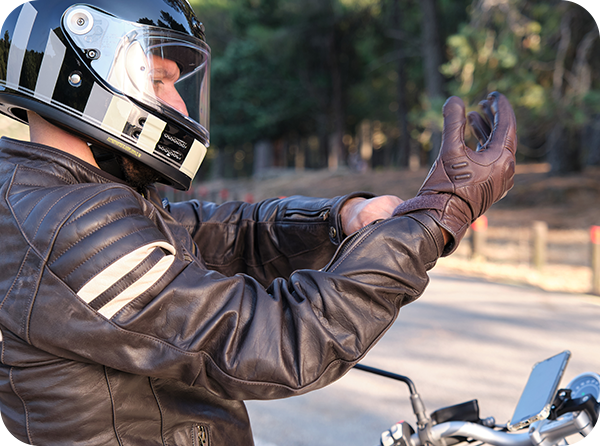 Photo of man in leather motorcycle coat and full face helmet putting on leather gloves with a motorcycle in the background