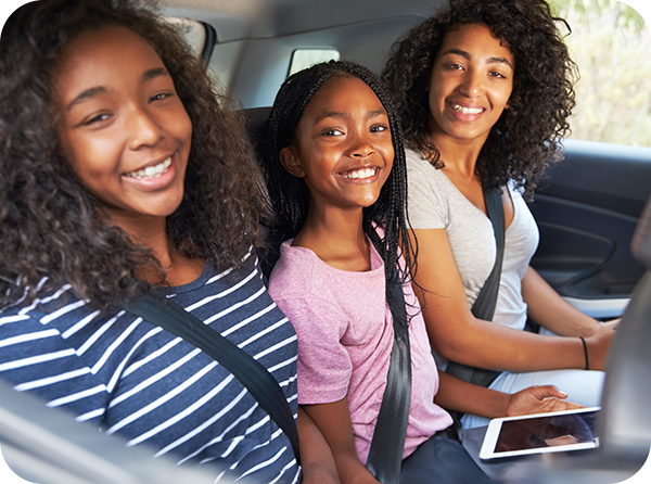 Photo of a teenager, pre-teen and mother in the backseat of a vehicle smiling with their seatbelts on