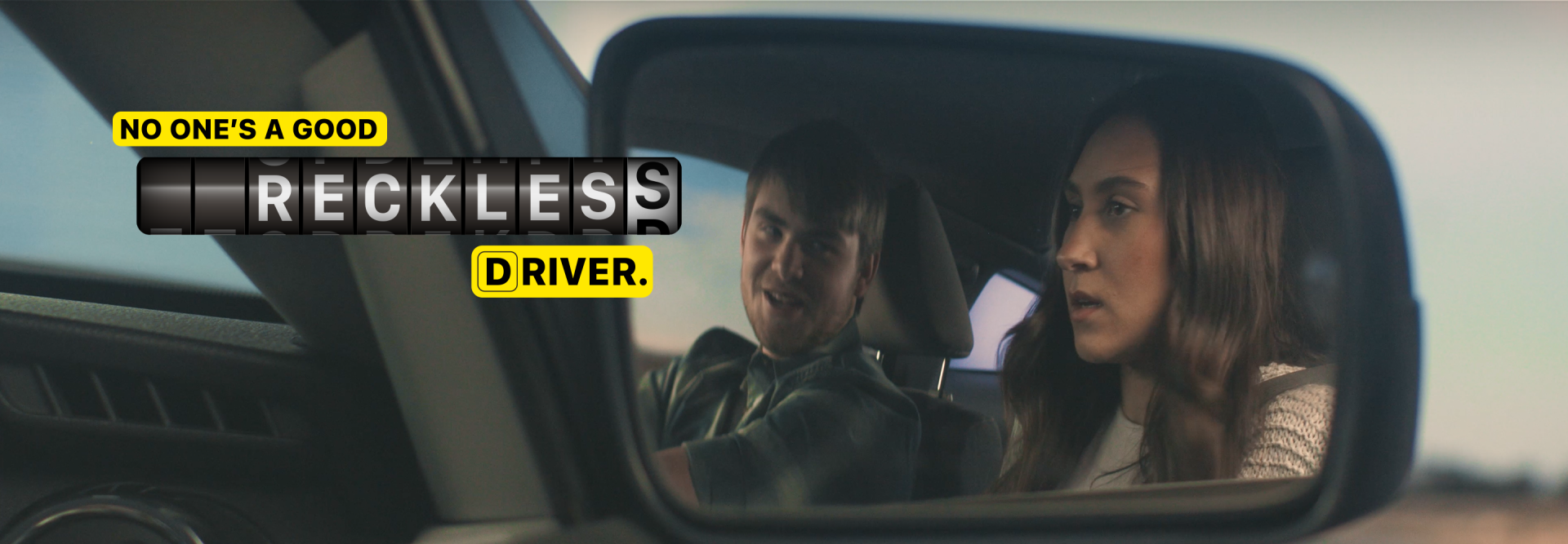 Photo of passenger's side rear view mirror showing reflection of a male driver talking and looking at a female in the passenger's seat that says "No one's a good reckless driver"