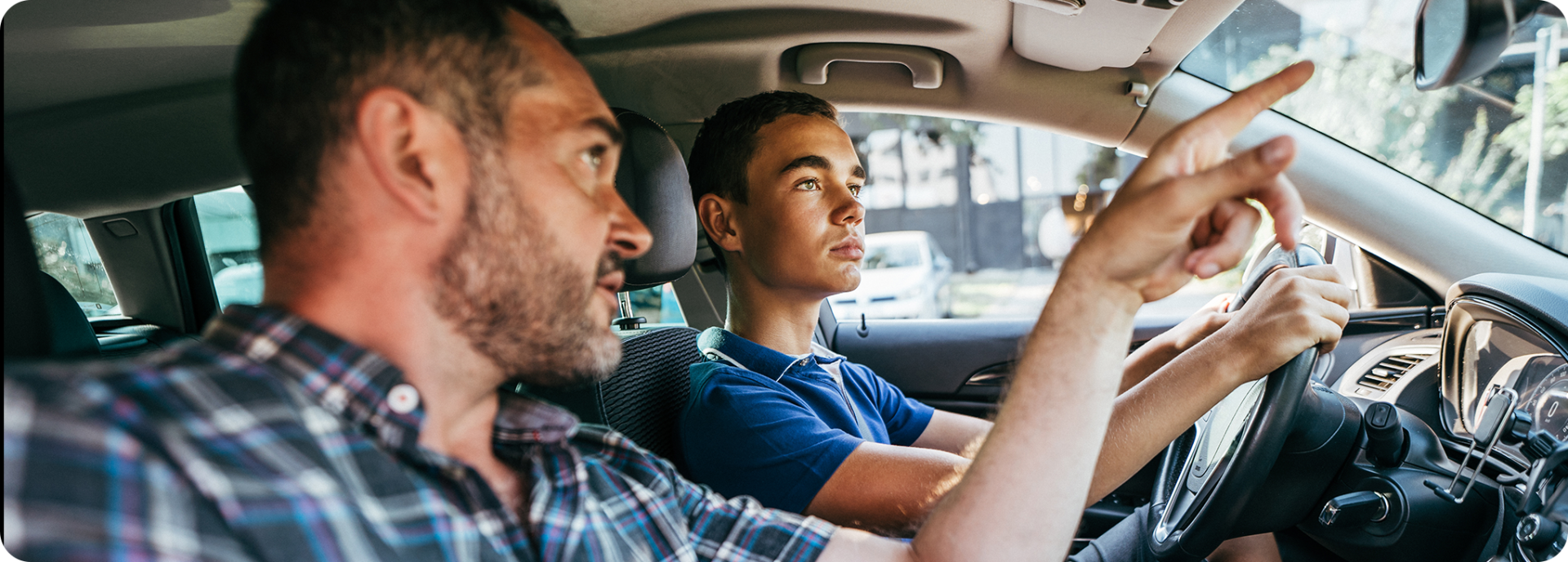 Photo of father sitting in the passenger's seat of a vehicle pointing to the rear view mirror as he presumably teaches his young son to drive