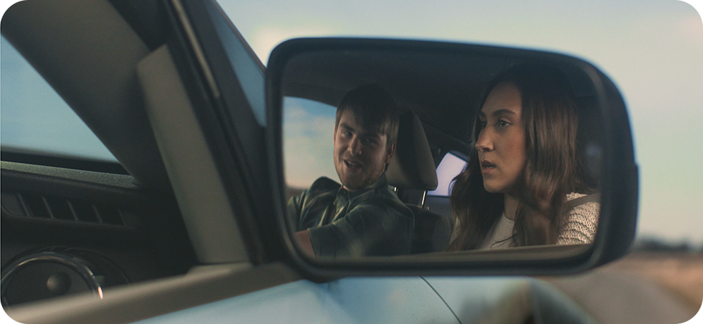 Photo of passenger's side rear view mirror showing reflection of a male driver talking and looking at a female in the passenger's seat who looks worried
