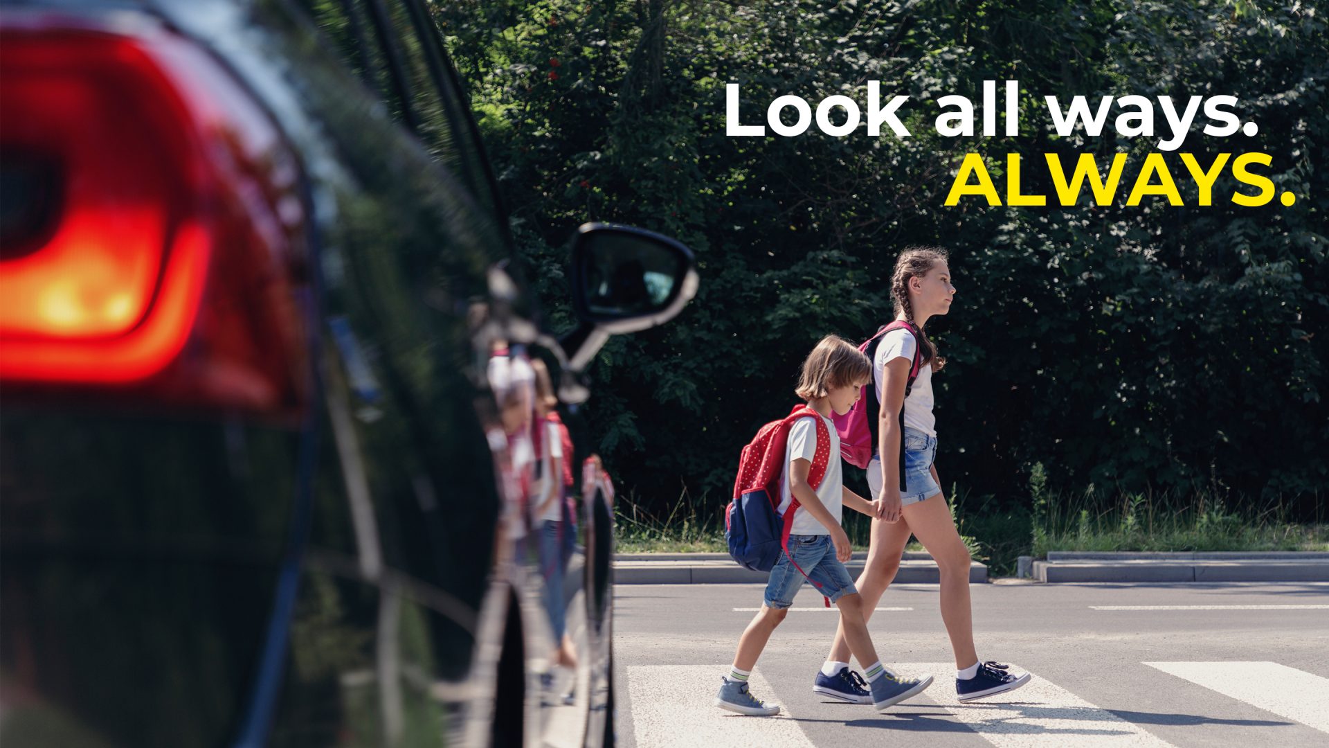 Photo of two young girls holding hands and walking across a while a car waits to go that says “Look all ways, always”